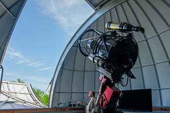A man stands inside near telescope looking outward.