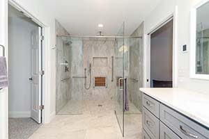 A newly renovated white bathroom with glass shower doors, gray cabinets and and marbled counter top.