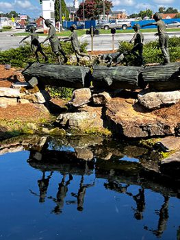 A sculpture of five kids waling across a fallen tree log and its reflection in the water below