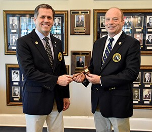 Incoming and Outgoing UYC Commodores dressed in navy jackets and khaki pants, facing camera perform symbolic handoff of the gavel.