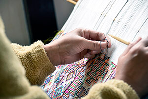 Closeup of a woman's hands holding thread and shuttle working on a pattern on hand loom.