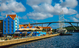 Downtown Willemstad waterfront with hotels on left, bridge in background, and dark blue water with fluffy white clouds in a blue sky.
