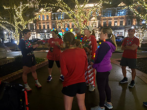 Members of Lanier Running Club gather on the Gainesville Square in the dark with Christmas lights in the background before Jingle Jog in December.