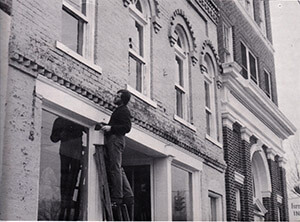 Black and white photo of Randy Wood standing on a ladder fixing the facade of a building on Main Street.
