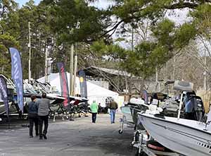 Scene from outdoor boat show with pine trees overhead, people walking and boats lining both sides of closed road.