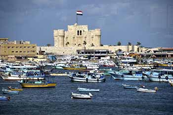 Photo of the Mediterranean with boats in front on blue water with a fortress in the background and the Egyptian flag flying.