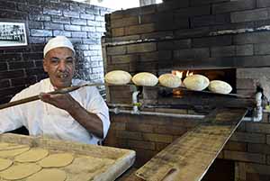 Photo of Egyptian baker holding his freshly cooked pita breads on a wooden board, just out of the oven in the background.