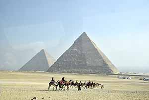 Photo of dessert, people on camels and the great Pyramids of Giza in the background.