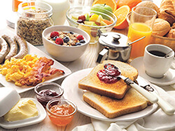 Closeup of table with white tablecloth and many breakfast items like eggs, bacon, sausage, toast, oatmeal, fruit and coffee - but no grits.