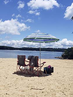 A sandy beach area with two chairs under an umbrella facing Lake Lanier.