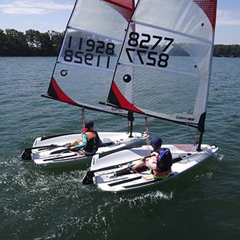 Two young students, each in their own sailboat, sail very close to each other. 