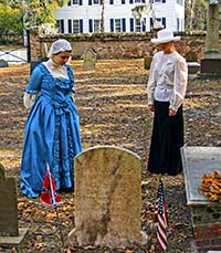Two young women dressed in period clothing standing in front of a tombstone showing respect for the dead.