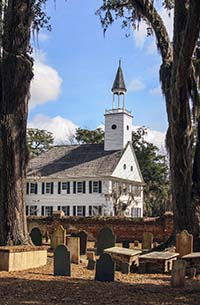 An old white church with mature oak trees on either side with the cemetery in foreground.