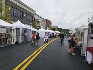 A view of street closed to car traffic lined with art booths and people walking through.