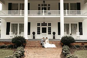 Front of large, 2-story white house, with brick steps with bride and groom sitting on the steps.