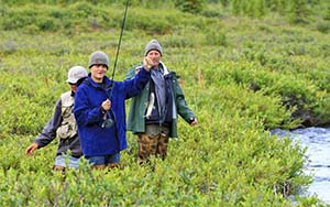 Three members of a family standing in tall grass next to a river with fishing poles.
