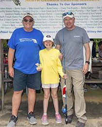 A smiling young girl in shorts, long sleeve shirt and cap, holds her fishing pole and trophy in hand with two men standing on either side of her sharing in her happy moment.