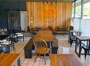 Inside view of restaurant 505 Eats. It has white tile flooring, wooden tables and chairs and a wood-panel wall with the 505 Eats logo.