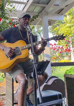 Guitar player, Tyrone Moultry, sitting on a wooden stool, playing guitar outside under a white, wooden gazebo.
