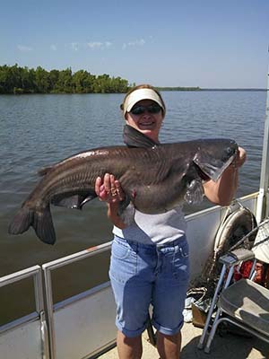 A woman in bluejean shorts, t-shirt and cap, holding a very large catfish in her hands. The lake and shoreline is in the background.