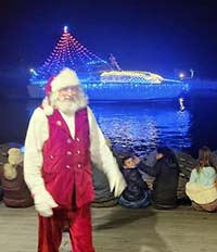 Nighttime picture with Santa on the dock with a boat dressed in holiday lights on Lake Lanier in the background.