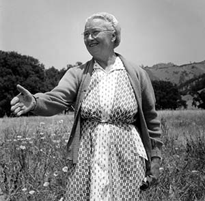 A copy of a black and white photo of a woman dressed in a dress and sweater standing in a field of grass and flowers with her hand outstretched to shake another person's hand. 