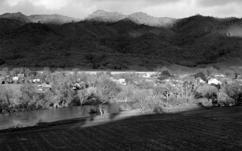A copy of a black and white photo taken by Pirkle Jones - of the Barryessa Valley, California in 1956 with mountains in the background, the town and riverbank.