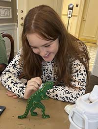 A Brenau student sitting at a desk painting a wooden dinosaur green.