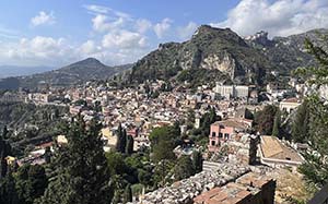 View of Taormina, Italy with rocky mountains in background, blue sky and buildings/homes made of rock, stone, brick in foreground.
