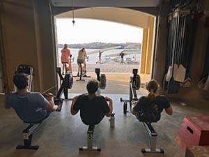A view of the back of three athletes on rowing machines, two on stationary bikes looking out from the boathouse to the lake.