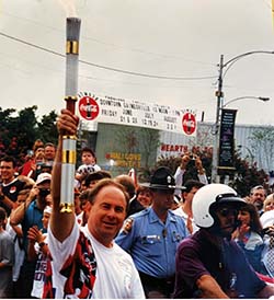 A photo from the 1996 torch relay, of Jim Mathis holding the torch in the foreground and many others in the background. 