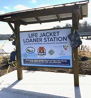 A Life Jacket Station at Lake Lanier Olympic Park. It has a sign held by two wooden posts with a slight roof and hooks on the posts that hold the life jackets. 