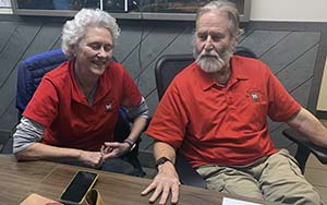 Janice (left) and John (right) Clark dressed in red golf shirts and khaki pants sitting at a desk. 