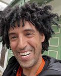 A head-shot of Abdu, the travel leader. He has dark skin, bright smile and curly dark hair, wearing a dark jacket with orange lanyard around his neck. 