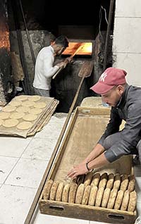 A photo of a community bread bakery showing one man putting bread into a large stone oven, and another man organizing the baked bread.