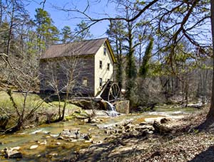 A side view of the old Sells Mill with a two story house and grist mill wheel with the river running over a shallow, rocky bottom.