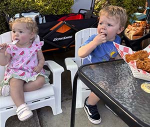 Two young toddlers sit in small chairs eating dinner. 